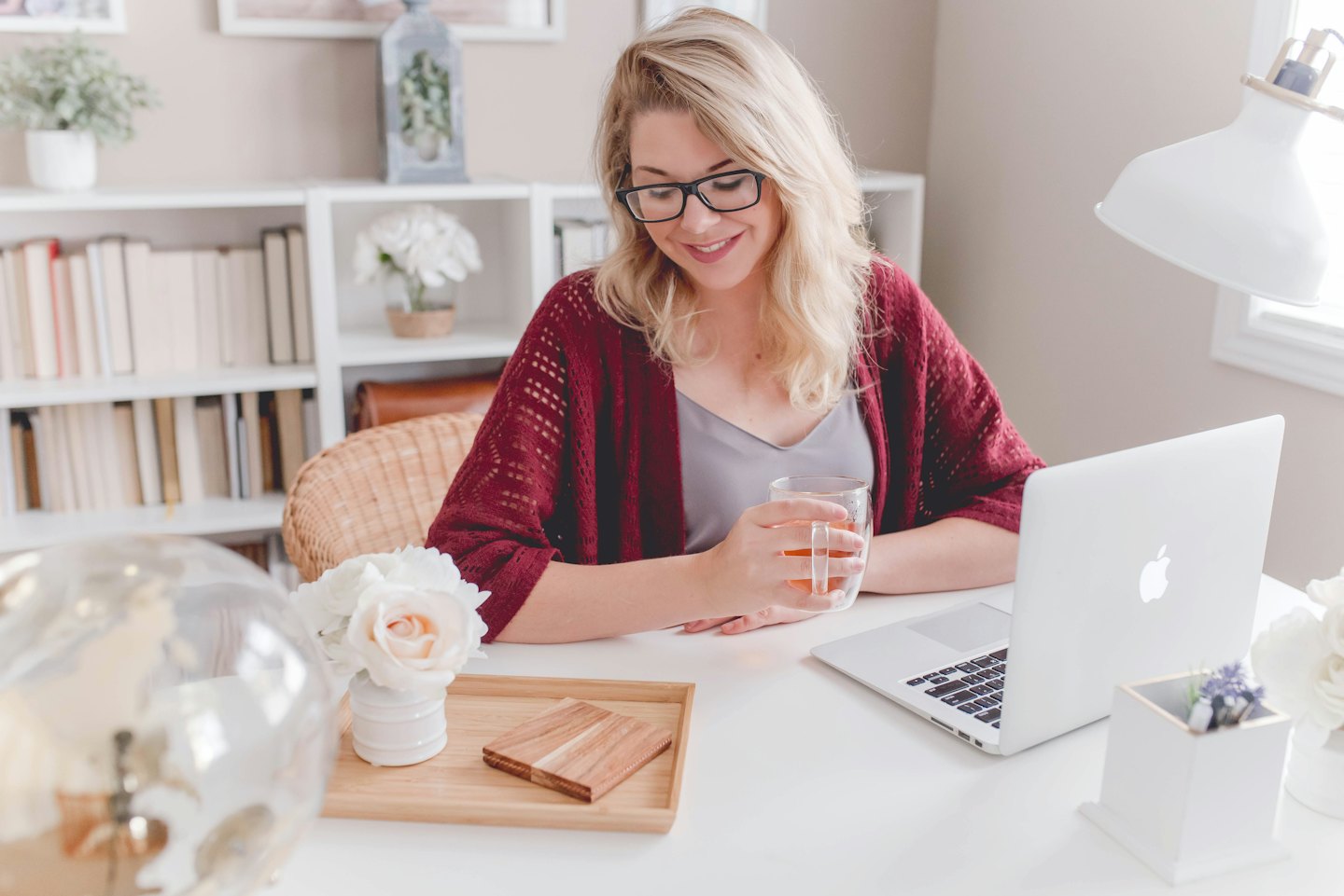 Woman in glasses smiles at laptop with tea at desk.