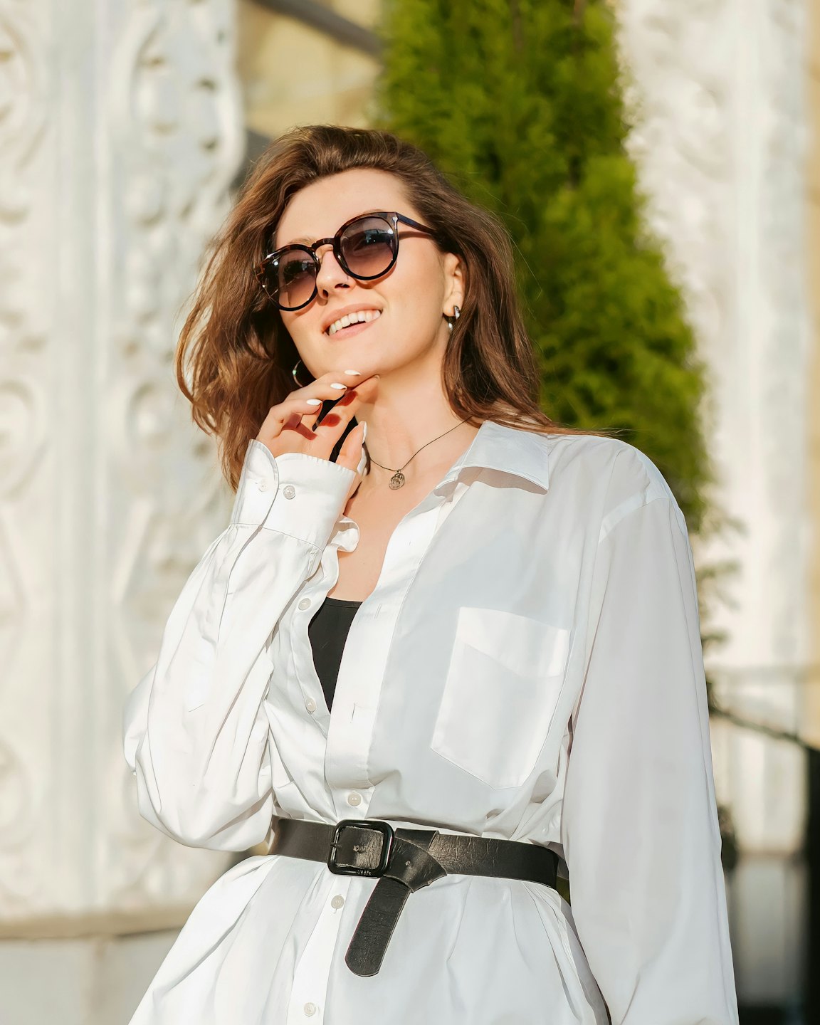 Woman wearing sunglasses and a white shirt touches her chin.